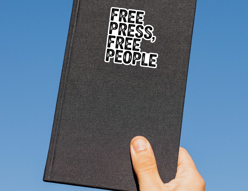May include: A black book with the words "FREE PRESS, FREE PEOPLE" in a white, bold font. The book is held by a hand against a clear blue sky.