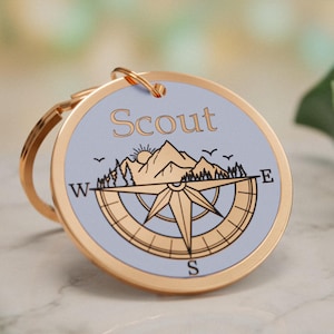 Könnte beinhalten: Ein runder Schlüsselanhänger aus goldfarbenem Metall mit einer hellblauen Einlage. Das Design zeigt das Wort Scout in schwarzer Schrift über einer schwarzen Strichzeichnung von Bergen, einer Sonne und einer Windrose mit den Buchstaben W, E und S.
