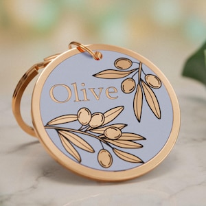Könnte beinhalten: Ein runder Schlüsselanhänger aus goldfarbenem Metall mit einer hellblauen Mitte. Das Design zeigt eine schwarz umrandete Illustration von Olivenzweigen mit Blättern und Früchten sowie das Wort Olive in einer goldfarbenen Serifenschrift.