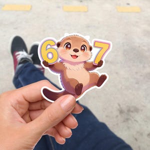 Könnte beinhalten: Ein Aufkleber mit einem Cartoon-Otter, der die Zahlen 6 und 7 hält. Der Otter ist braun und hellbraun mit einem lächelnden Gesicht. Die Zahlen sind gelb mit weißem Rand. Der Aufkleber wird in einer Hand gehalten.