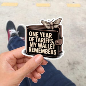 Könnte beinhalten: Ein Aufkleber mit einer braunen Geldbörse und dem Text "ONE YEAR OF TARIFFS, MY WALLET REMEMBERS". Eine Mottenillustration befindet sich über der Geldbörse. Der Aufkleber wird von einer Person in der Hand gehalten, mit einem verschwommenen Hintergrund von Beinen und Schuhen.