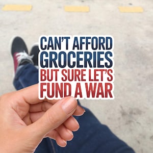 Könnte beinhalten: Ein weißer Aufkleber mit dem Text "CAN'T AFFORD GROCERIES BUT SURE LET'S FUND A WAR" in Blau und Rot. Der Aufkleber wird in einer Hand gehalten, mit einem verschwommenen Hintergrund aus Jeans und Turnschuhen.