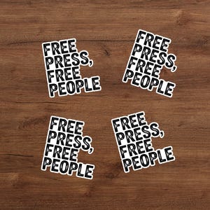 May include: Four white stickers with the text "FREE PRESS, FREE PEOPLE" in a black, bold font. The stickers are arranged on a wooden surface. The text is stacked vertically, with a comma after "PRESS".
