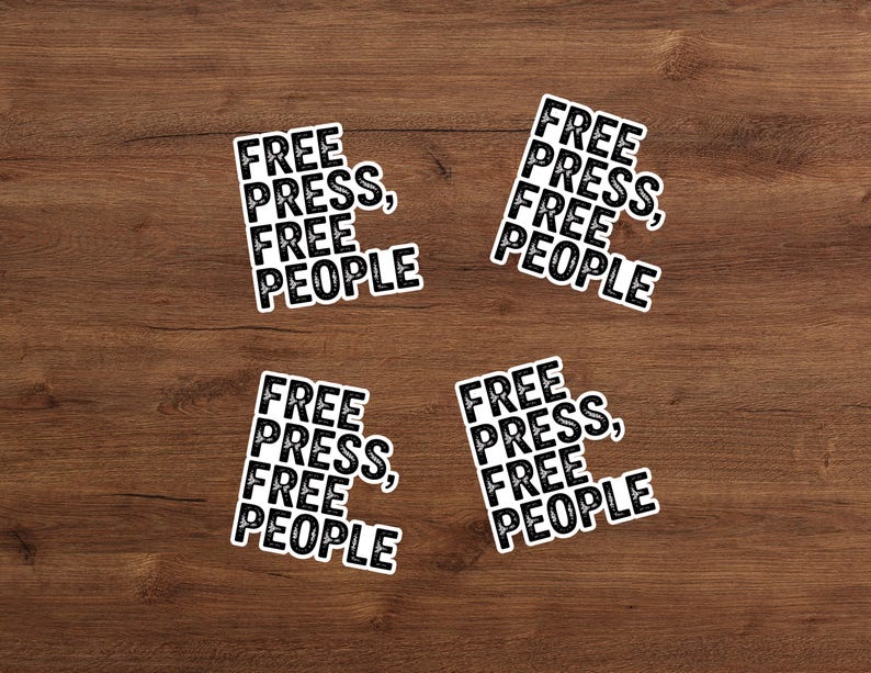 May include: Four white stickers with the text "FREE PRESS, FREE PEOPLE" in a black, bold font. The stickers are arranged on a wooden surface. The text is stacked vertically, with a comma after "PRESS".