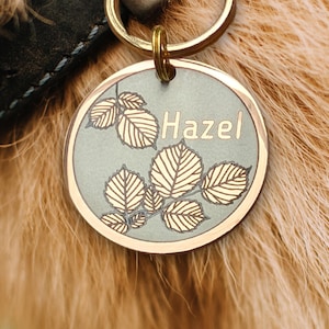 Może przedstawiać: Okrągły, srebrny identyfikator dla psa z pozłacanym rantem i wzorem liści. Identyfikator ma wygrawerowane imię "Hazel".