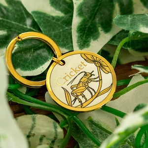 Könnte beinhalten: Goldfarbener Schlüsselanhänger aus Metall mit einem runden Anhänger, der eine detaillierte Illustration eines Grillen auf einer Blume zeigt. Der Anhänger ist mit dem Wort "Cricket" graviert.
