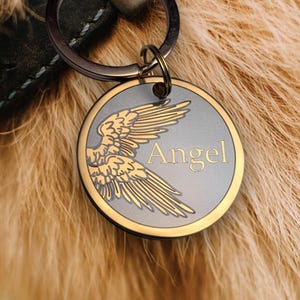 May include: A round, metallic pet tag with a gold-colored wing design and the word "Angel" in gold lettering. The tag is attached to a key ring and a black leather collar. The background is the light brown fur of a dog.