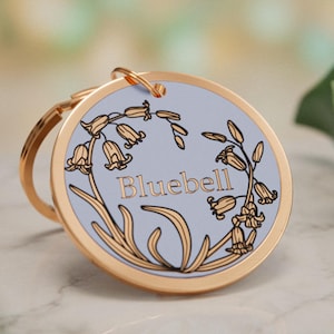Könnte beinhalten: Ein runder Schlüsselanhänger aus goldfarbenem Metall mit einer hellblauen Einlage in der Mitte. Das Design zeigt eine schwarze Strichzeichnung von Glockenblumen, die das Wort Bluebell in goldener Schrift umrahmen. Ein goldener Ring ist oben befestigt.