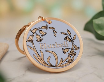 May include: A round gold-toned metal keyring featuring a light blue centre inlay. The design displays a black line art illustration of bluebell flowers surrounding the word Bluebell in gold lettering. A gold metal ring is attached to the top.