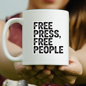 May include: White ceramic mug with the text "FREE PRESS, FREE PEOPLE" in bold black letters. The mug has a light blue interior and a white handle. The mug is held in front of a blurred background.