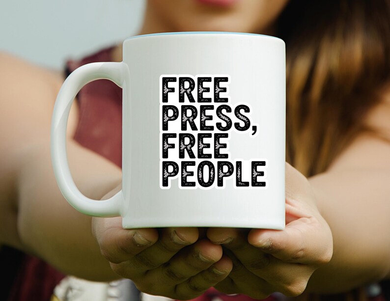 May include: White ceramic mug with the text "FREE PRESS, FREE PEOPLE" in bold black letters. The mug has a light blue interior and a white handle. The mug is held in front of a blurred background.