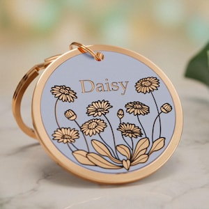 May include: A round gold-toned metal keychain featuring a light blue center inlay. The inlay displays a black line-art illustration of daisy flowers and the name Daisy printed in a gold-colored serif font at the top center of the circular disc.
