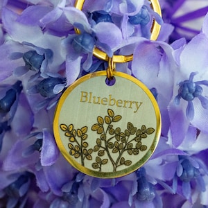 Könnte beinhalten: Ein goldfarbener Metallschild mit dem Wort "Blueberry" und einem stilisierten Bild von Blaubeerblättern. Das Schild hängt an einem Schlüsselring und ist von lila Blüten umgeben.