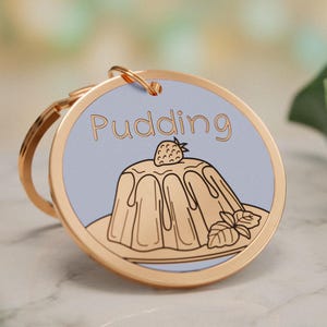 Könnte beinhalten: Ein runder Schlüsselanhänger aus goldfarbenem Metall mit hellblauem Hintergrund und einer Strichzeichnung eines Pudding-Desserts, das mit einer Erdbeere und Minzblättern garniert ist. Das Wort Pudding steht in einfacher brauner Schrift darüber.