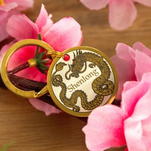 Könnte beinhalten: Goldfarbener Schlüsselanhänger mit einem runden Anhänger, der eine detaillierte Illustration eines Drachen mit dem Wort "Shenlong" zeigt. Der Schlüsselanhänger ist von rosa Stoffblumen umgeben.