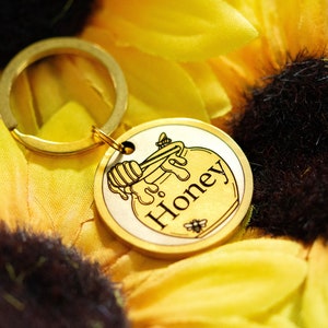 Op de afbeelding: Een goudkleurige sleutelhanger met een ronde bedel met een honingpotontwerp en het woord "Honey". De bedel is bevestigd aan een gouden ring. De achtergrond is een gele zonnebloem.