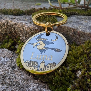 Op de afbeelding: Een gouden metalen hondenpenning met een blauwe achtergrond waarop een heks op een bezem vliegt. De penning heeft de naam "Luna" gegraveerd.