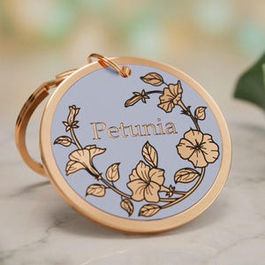 Könnte beinhalten: Eine runde Haustiermarke aus goldfarbenem Metall mit einer hellblauen Mitte. Die Marke zeigt den Namen Petunia in goldener Schrift, umgeben von einer goldenen Blumenranken-Illustration. Ein goldener Metallring ist oben an der Marke befestigt.