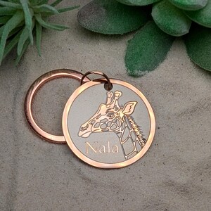 May include: A round, copper-colored pet tag with a giraffe head engraving and the name "Nala" in a serif font. The tag is attached to a copper-colored split ring. The background is a sandy texture.