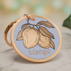 May include: A round gold-toned metal keychain featuring a light blue circular insert. The insert displays a black line-art illustration of two mangoes on a branch with leaves and the word Mango printed in a simple serif font below the fruit.
