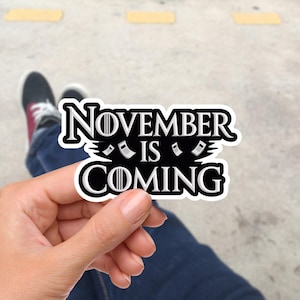 Può includere: Un adesivo in bianco e nero con la scritta "NOVEMBER IS COMING". L'adesivo ha un bordo bianco ed è tenuto in mano. Lo sfondo è sfocato, mostrando jeans e scarpe da ginnastica.