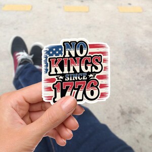 Könnte beinhalten: Ein rechteckiger Aufkleber mit patriotischem Design. Der Aufkleber zeigt den Text "NO KINGS SINCE 1776" in fetten, umrandeten Buchstaben, vor dem Hintergrund der amerikanischen Flagge. Der obere Teil zeigt das blaue Sternenfeld, während der untere Teil rote und weiße Streifen hat.