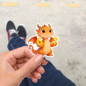 Könnte beinhalten: Ein Aufkleber mit einem orangefarbenen Cartoon-Drachen, der die Zahlen 6 und 7 hält. Der Drache hat kleine Hörner, Flügel und einen freundlichen Gesichtsausdruck. Der Aufkleber wird in einer Hand gehalten, mit einem verschwommenen Hintergrund aus Beinen und einer Betonoberfläche.