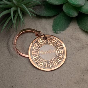 Könnte beinhalten: Ein roségoldener Schlüsselanhänger mit einem kreisförmigen Anhänger. Der Anhänger zeigt den Namen "Simba" in der Mitte, umgeben von einem komplizierten geometrischen Muster. Der Schlüsselanhänger liegt auf einer sandigen Oberfläche, mit grünen Pflanzen im Hintergrund.