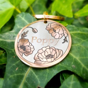 Pu&ograve; includere: Un medaglietta rotonda per animali domestici con bordo dorato e il nome "Poppy" inciso, circondato da disegni di fiori di papavero. Un anello dorato &egrave; presente per l'attacco. Lo sfondo &egrave; composto da foglie verdi.