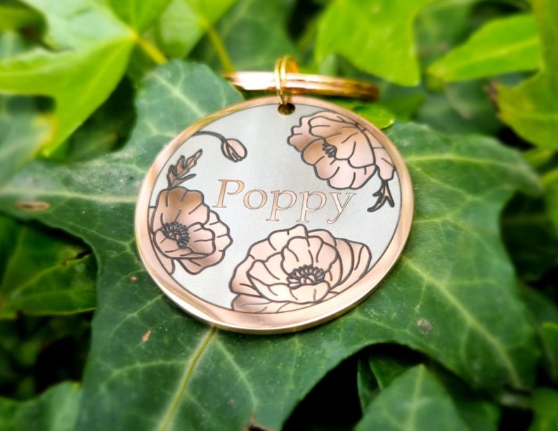 Pu&ograve; includere: Un medaglietta rotonda per animali domestici con bordo dorato e il nome "Poppy" inciso, circondato da disegni di fiori di papavero. Un anello dorato &egrave; presente per l'attacco. Lo sfondo &egrave; composto da foglie verdi.