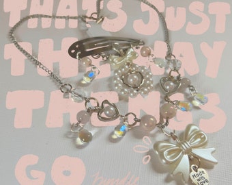 May include: A silver chain necklace with a silver heart charm, pearl beads, and a silver bow charm. The necklace also has a silver hair clip with a pearl and bow charm. The charms are attached to the chain with clear crystal beads. The necklace is on a pink background with the text "That's just the way things go" written in pink.