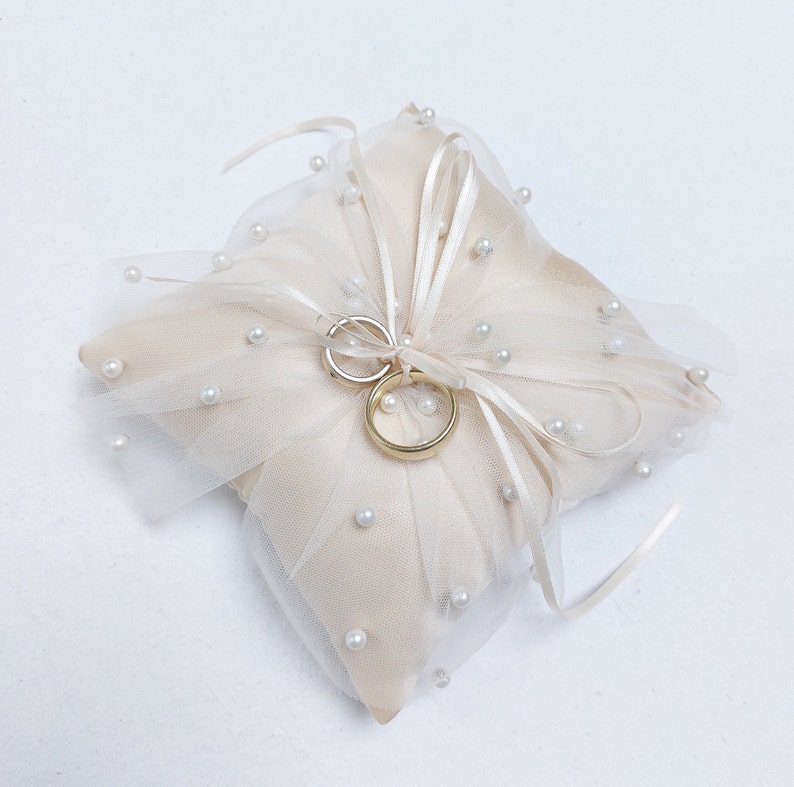 K&ouml;nnte beinhalten: Ein wei&szlig;es Satin-Kissen mit T&uuml;ll&uuml;berzug und Perlenbesatz. Zwei goldene Eheringe liegen auf dem Kissen.