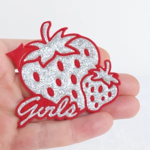 May include: Red and silver glitter iron-on patch with two strawberries and the word "Girls" in a cursive font.