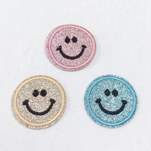 May include: Three sparkly smiley face iron-on patches in pink, gold, and blue.