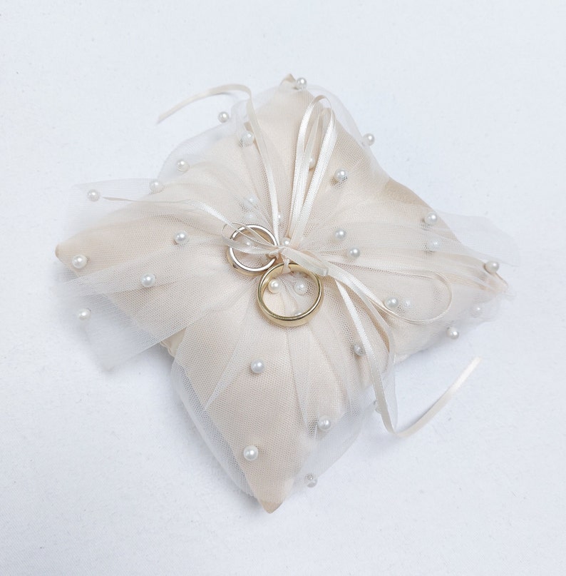 K&ouml;nnte beinhalten: Ein wei&szlig;es Satin- und T&uuml;ll-Ringkissen mit Perlenbesatz und zwei goldenen Eheringen.
