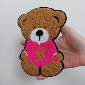 Könnte beinhalten: Ein brauner Teddybär, der einen rosa Overall mit einem goldenen Buchstaben "P" auf der Vorderseite trägt. Der Bär sitzt mit gekreuzten Beinen und hat ein weißes Gesicht mit schwarzen Augen und einer schwarzen Nase.