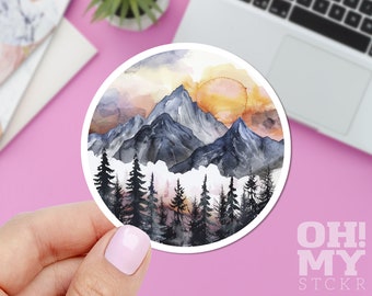 May include: A round sticker with a watercolor painting of a mountain range with a sunset in the background. The mountains are blue and gray, and the sunset is orange and yellow. There are trees in the foreground.