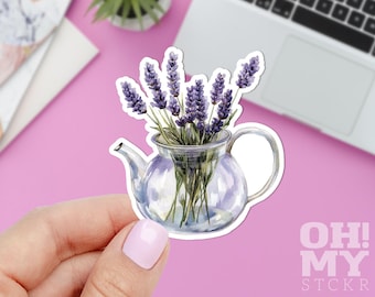 Puede incluir: Una pegatina con una ilustración de acuarela de una tetera transparente llena de ramitas de lavanda. La lavanda tiene flores moradas y tallos verdes. La pegatina tiene un borde blanco y el texto "OH MY STCKR" en rosa.