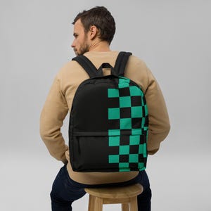 Puede incluir: Mochila negra con un diseño a cuadros negro y verde azulado en el lado derecho. La mochila tiene correas negras para los hombros y un bolsillo frontal. La persona de la imagen lleva la mochila.