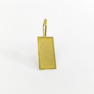 Pode incluir: Uma etiqueta retangular dourada com uma superfície texturizada e uma alça superior curva. A etiqueta é feita de metal e está sobre um fundo branco. A etiqueta é provavelmente para um porta-chaves ou bagagem.