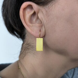 Pode incluir: Um brinco retangular dourado com superfície texturizada, pendurado em um gancho dourado. O brinco é usado na orelha de uma pessoa com cabelo escuro e um colar de ouro.