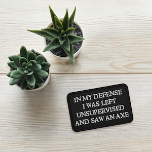 May include: Black patch with white text that reads "In my defense I was left unsupervised and saw an axe". The patch is on a white wooden surface with two small green plants in white pots.