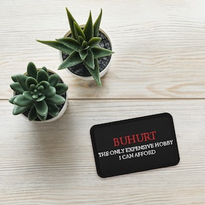 May include: Black patch with white text that reads "BUH HURT THE ONLY EXPENSIVE HOBBY I CAN AFFORD". The text is in red. The patch is on a light wood surface with two small green plants in white pots.