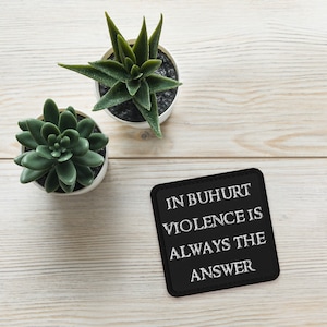 May include: A black square patch with white embroidered text that reads "In Buhurt Violence Is Always The Answer" on a white wooden surface with two green succulent plants in white pots.