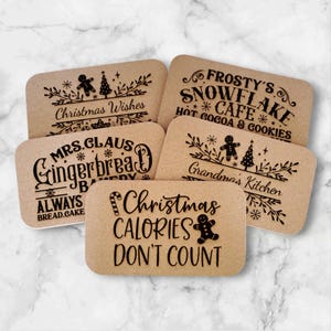 May include: Set of six brown wood coasters with black laser-engraved Christmas-themed designs. The designs include "Christmas Wishes", "Frosty's Snowflake Cafe", "Mrs. Claus Gingerbread Bakery", "Grandma's Kitchen", "Always Bread.Cake", and "Christmas Calories Don't Count".