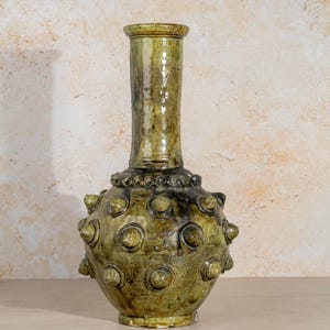 Könnte beinhalten: Eine hohe, bauchige Keramikvase mit glänzender olivgrüner Glasur. Der kugelförmige Körper weist ein sich wiederholendes Muster aus erhabenen, kreisförmigen Noppen oder Spitzen auf. Sie hat einen schmalen, zylindrischen Hals mit einem ausgestellten Rand.