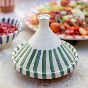 May include: A white ceramic tagine lid with green stripes. The lid is cone-shaped and has a small knob on top.