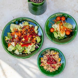 May include: Three green ceramic plates with different salads. The plates have a raised rim and a textured surface. The salads include a green salad with carrots, corn, and red cabbage, a green salad with tomatoes and carrots, and a couscous salad with pomegranate seeds.