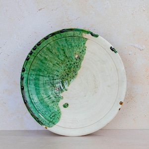 May include: A handmade ceramic plate with a white and green glaze. The plate is round, with a half-green, half-white design. The green glaze has a textured, organic appearance, and the rim is decorated with black dots.