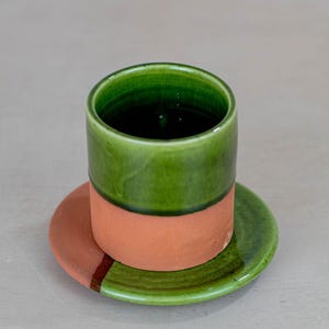May include: A handmade ceramic cup and saucer set. The cup is green on top and terracotta on the bottom. The saucer is green with a terracotta stripe. The set is designed for serving beverages.